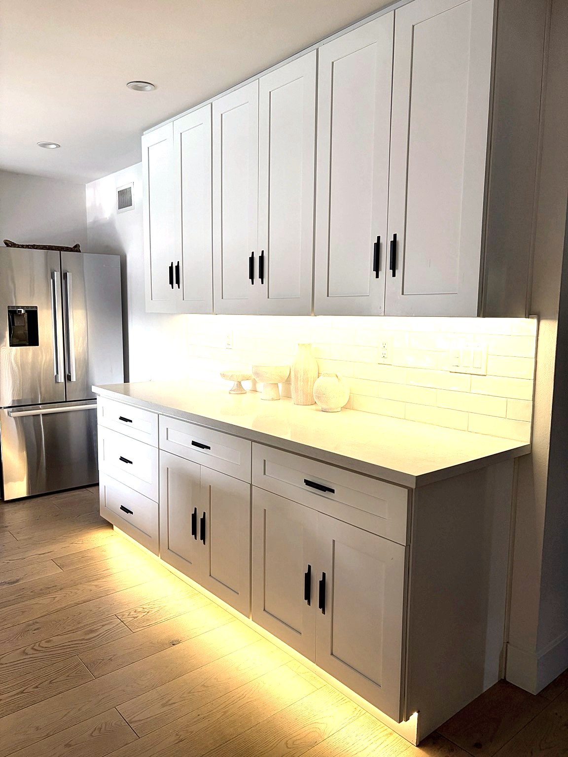 White shaker kitchen with black hardware, glowing under-cabinet and toe-kick lighting and stone vessels