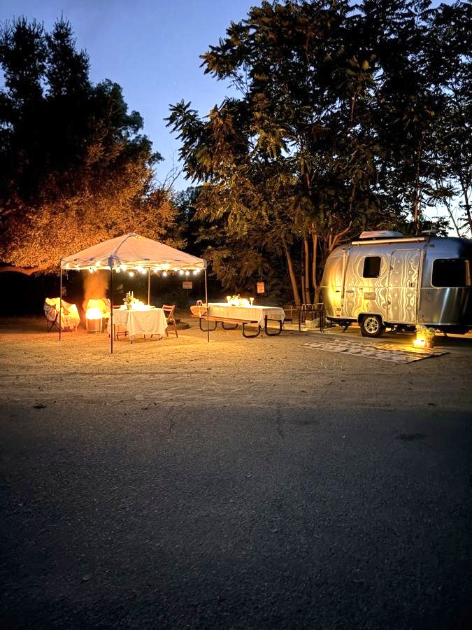 Campsite at dusk with a string-lit canopy, dressed tables and a glowing Airstream