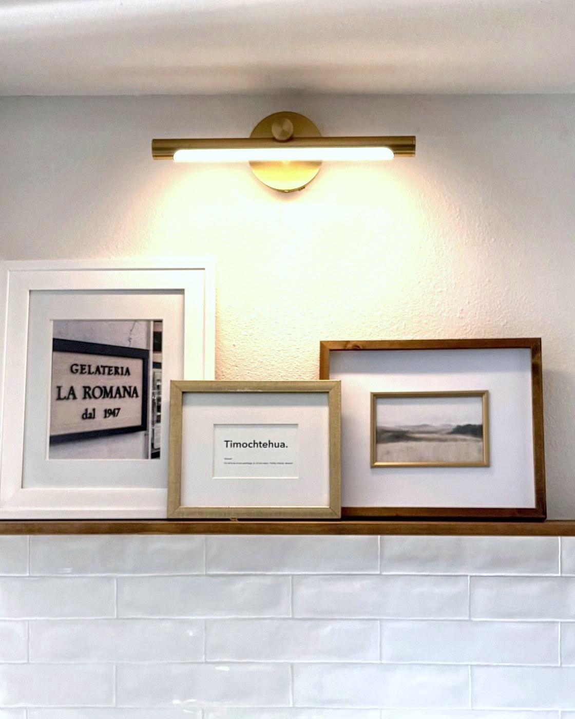 Floating walnut shelf with layered white, gold and wood frames under a brass picture light, above handmade white tile